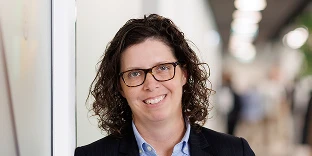 A woman with curly dark hair and glasses, smiling while wearing a dark pinstripe blazer over a light blue shirt. She is leaning against a white wall with a blurred office hallway in the background.
