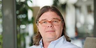 A man with shoulder-length light brown hair and orange-rimmed glasses, wearing a light blue collared shirt. He has a calm expression and is posed in a bright office with blurred greenery in the background.