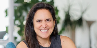 A woman with long dark hair and a wide, joyful smile. She is wearing a grey sleeveless top and a necklace, posed in front of a bright background with blurred green houseplants.