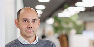 A man with a shaved head and thin, rimless glasses, wearing a grey sweater over a white collared shirt. He has a neutral expression and is leaning against a white wall in a modern office with blurred hallway lights in the background.