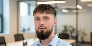 A man with short brown hair and a thick, well-groomed beard, wearing a light blue collared shirt. He has a neutral expression and is posed in a modern, bright office with blurred desks and windows in the background.