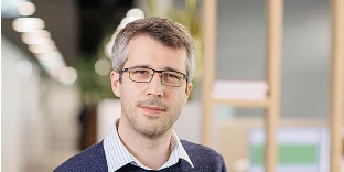 A man with short, salt-and-pepper hair and dark-rimmed glasses, wearing a navy blue sweater over a blue and white striped shirt. He has a calm expression and is posed in a bright, modern office with blurred shelving in the background.
