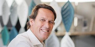 A man with brown hair and an approachable, friendly expression, wearing a white button-down shirt in a bright, modern office.