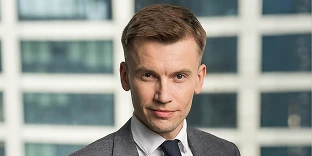 A man with short brown hair and a professional expression, wearing a grey suit jacket, white shirt, and navy blue tie. He is posed in front of a blurred background showing the windows of a high-rise office building.