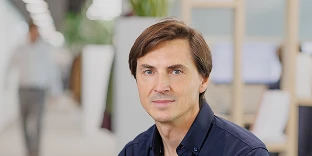 A man with short brown hair swept to the side and a calm, professional expression, wearing a navy blue button-down shirt. He is posed in a modern office with blurred desks, computers, and a hallway in the background.