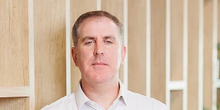 A man with short brown hair and blue eyes, wearing a light-colored button-down shirt. He has a calm, professional expression and is posed against a background of vertical wooden slats.