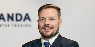 A man with short brown hair and a beard, smiling while wearing a dark suit and a blue tie. He is posed in front of a white wall featuring a blurred company logo.