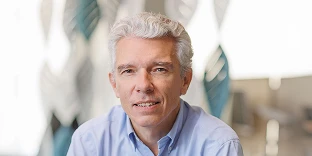 A man with wavy white hair and a professional expression, wearing a light blue button-down shirt. He is posed in a bright office environment with blurred leaf-shaped panels in the background.