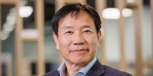 A man with short black hair and a kind, subtle smile, wearing a dark textured blazer over a light blue pinstripe shirt. He is posed in a modern office with blurred wooden architectural elements in the background.