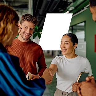 A diverse group of colleagues smiling and shaking hands in a modern office hallway. A woman in a white t-shirt shakes hands with a coworker while a man in an orange sweater looks on encouragingly.
