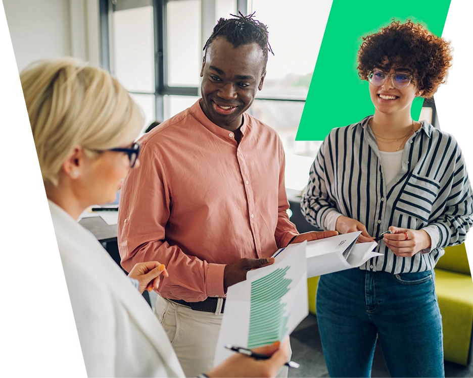 A diverse group of three colleagues smiling and collaborating in a bright office. They are reviewing printed documents and data charts together during a casual meeting.