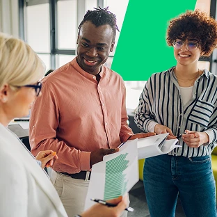 A diverse group of three colleagues smiling and collaborating in a bright office. They are reviewing printed documents and data charts together during a casual meeting.