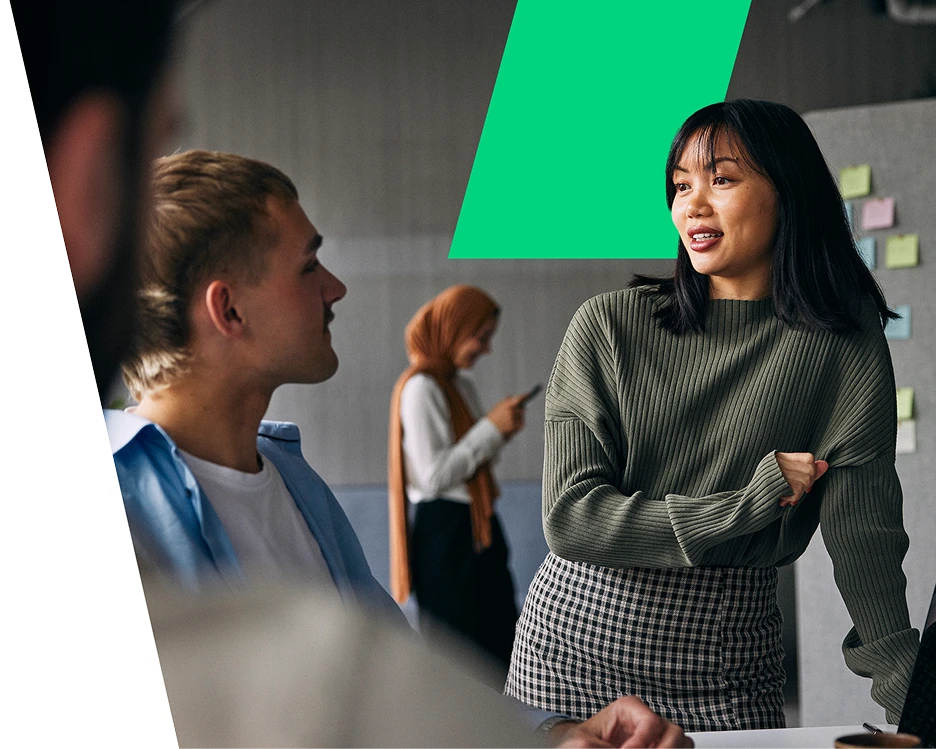 An East Asian woman with straight black hair and a green ribbed sweater stands and speaks during a collaborative team meeting in a modern office. She is looking toward a male colleague sitting at the desk who is listening intently. In the blurred backgrou